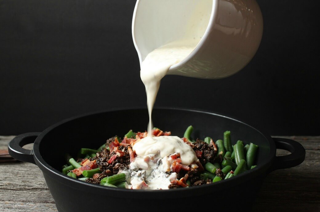 White sauce being poured over a black casserole dish filled with bacon, mushrooms, and green beans. 