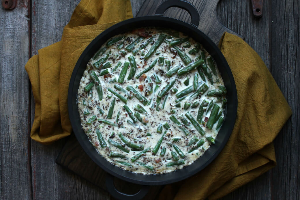 Overhead view of fresh green beans, bacon, mushrooms, and cream sauce in a black dish on a wooden surface.