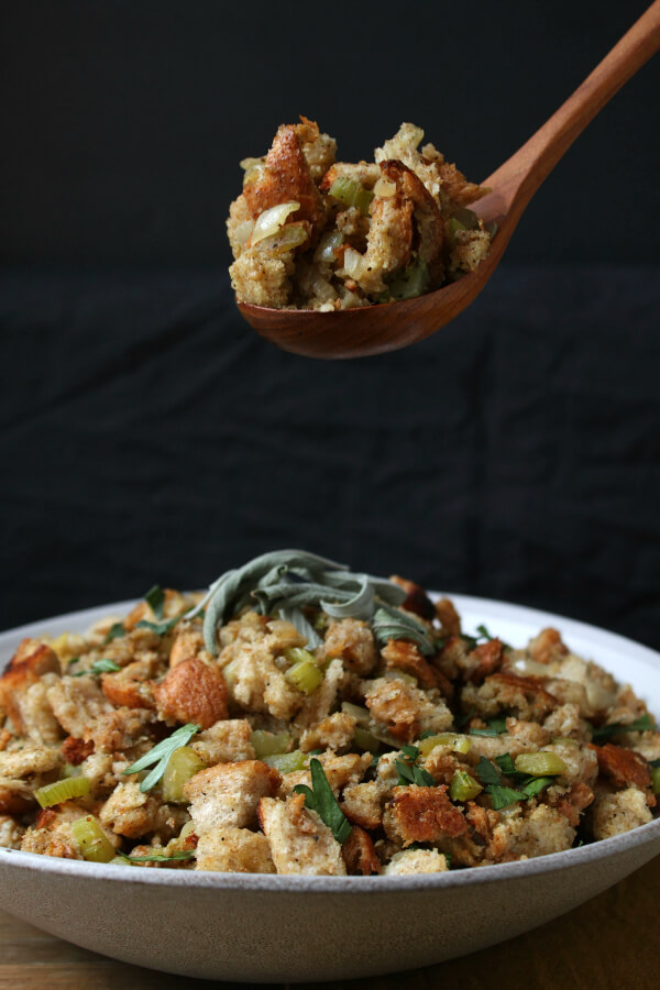 A spoonful of Homemade Stuffing above a grey bowl filled with classic bread stuffing studded with onions and celery.