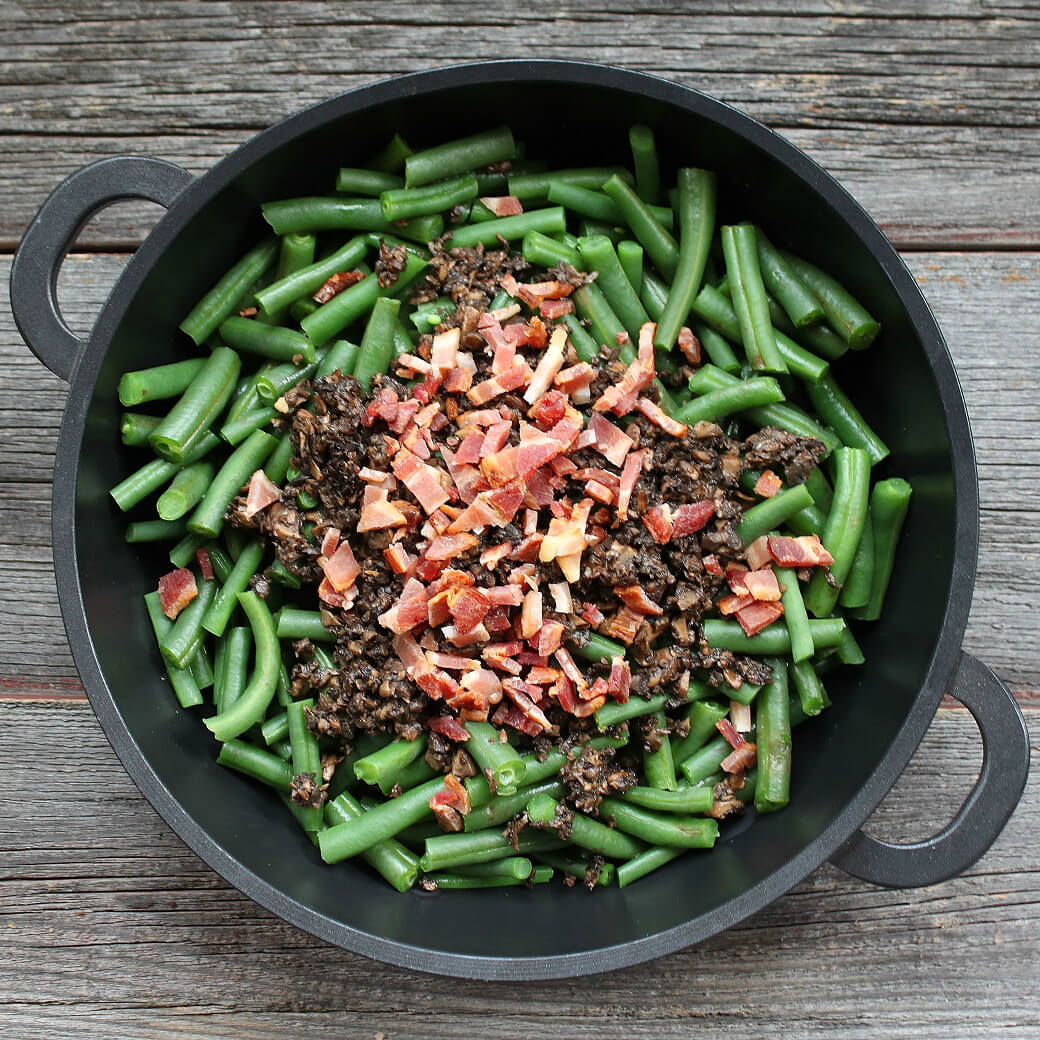 Overhead view of fresh green beans, mushrooms, and bacon in a black dish on a wooden surface.