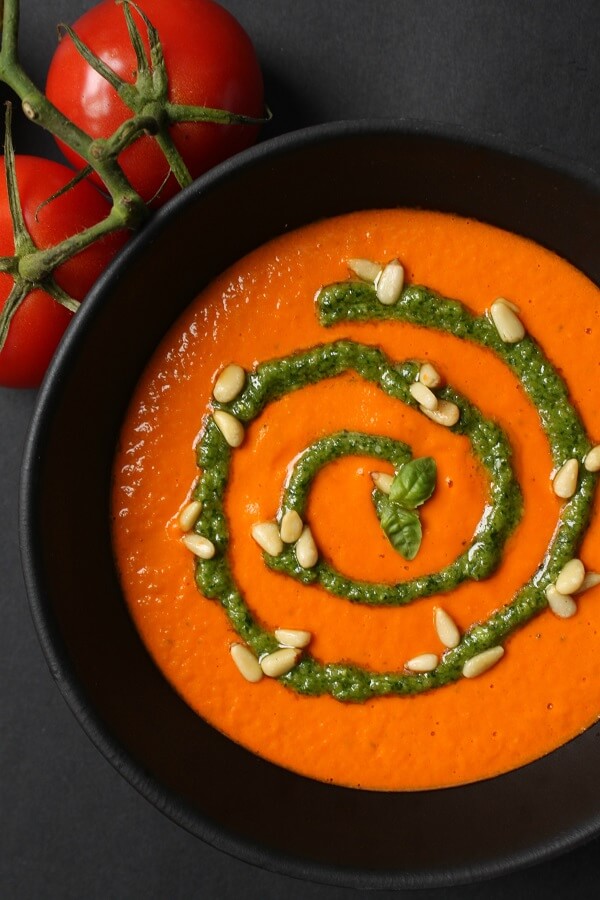 A black bowl of bright orange creamy tomato soup with a swirl of green pesto and scattered pine nuts.