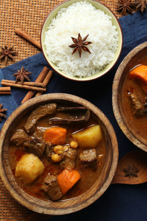 Massaman Beef Coconut Curry - Wooden bowls filled with bites of potato, sweet potato, and beef in a rich sauce. Surrounded by a small bowl of rice and spices.