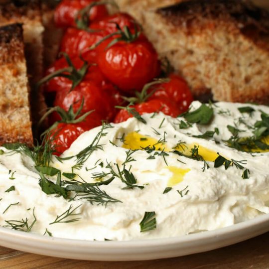 Whipped Feta - Roasted tomatoes and dark sliced bread surround a mound of creamy feta topped with olive oil and fresh green herbs.