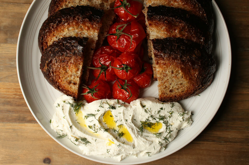 Whipped Feta - Roasted tomatoes and dark sliced bread surround a mound of creamy feta topped with olive oil and fresh green herbs.