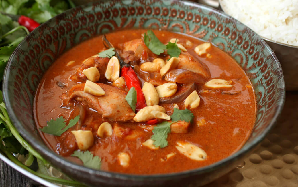 Peanut Butter Chicken Curry - Side view of an ornate bowl containing dark red curry with chicken, onions, red chili pepper, peanuts, and cilantro.