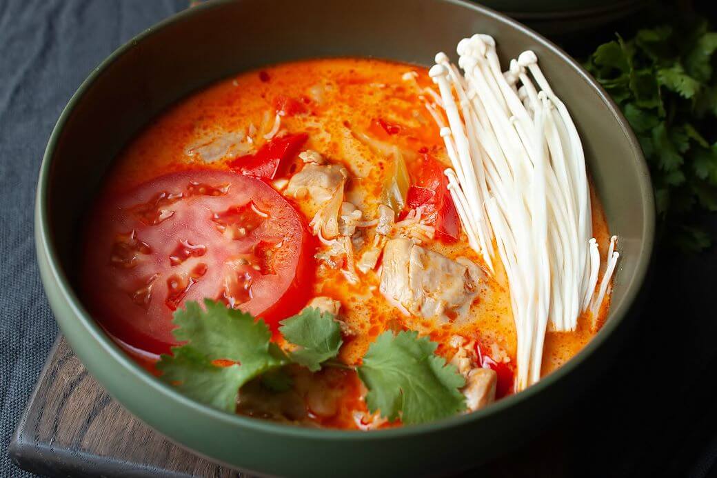 Thai Red Curry Chicken Soup - a green bowl of soup with creamy red broth, tomato, chicken, peppers, cilantro, and enoki mushrooms.