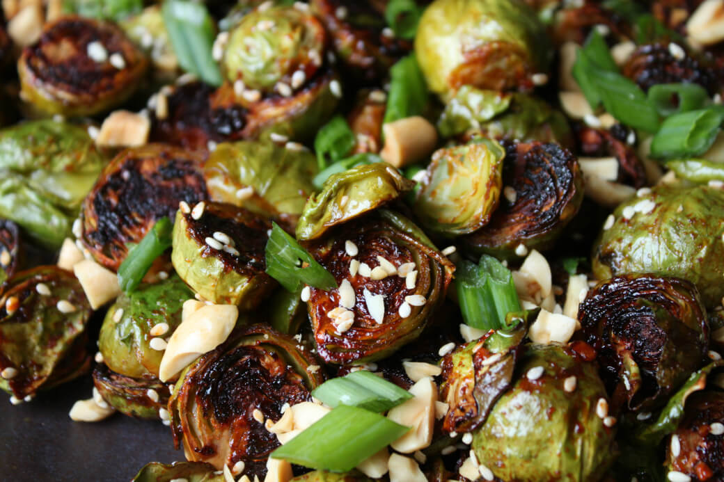 Maple Gochujang Roasted Brussels Sprouts - Dark roasted Brussels sprouts topped with sesame seeds, peanuts, and green onions on a black platter.