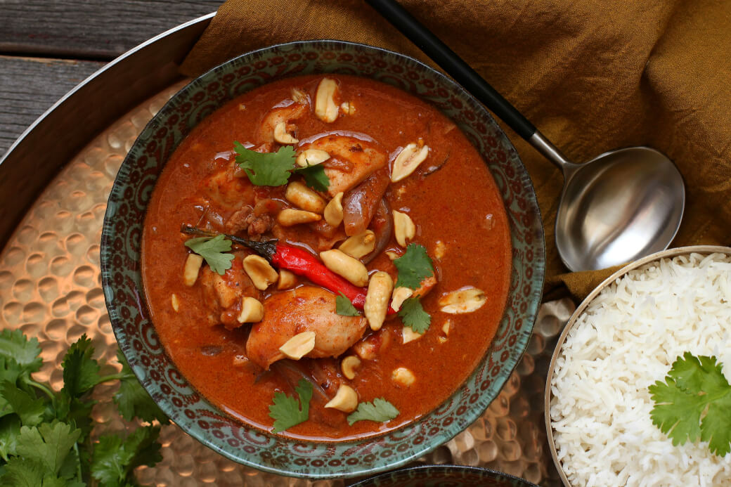 Peanut Butter Chicken Curry - One ornate bowl containing dark red curry with chicken, onions, red chili pepper, peanuts, and cilantro.