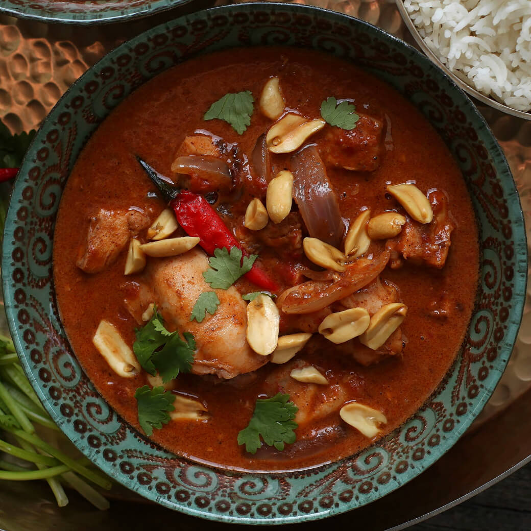 Peanut Butter Chicken Curry - Close up of an ornate bowl containing dark red curry with chicken, onions, red chili pepper, peanuts, and cilantro.