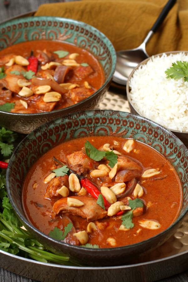 Peanut Butter Chicken Curry - Two ornate bowls containing dark red curry with chicken, onions, red chili pepper, peanuts, and cilantro.