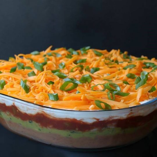 7 Layer Dip - A glass dish of layered dip topped with shredded cheese and sliced green onions.