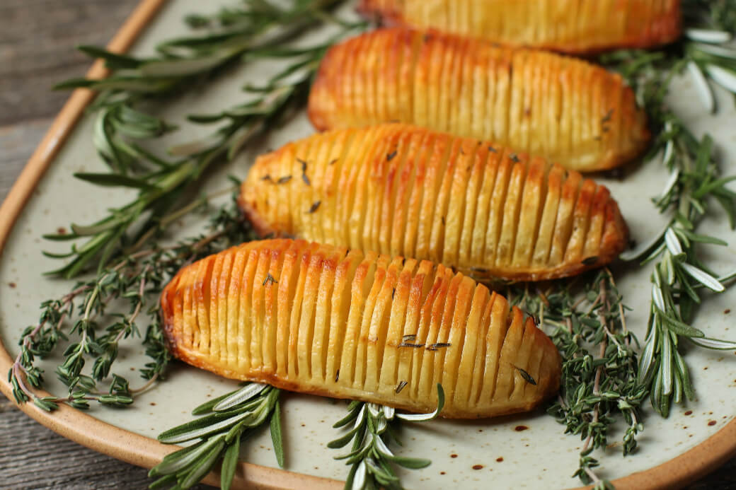 Crispy Herbed Hasselback Potatoes - Close up of crispy golden potatoes rest on a bed of fresh herbs.