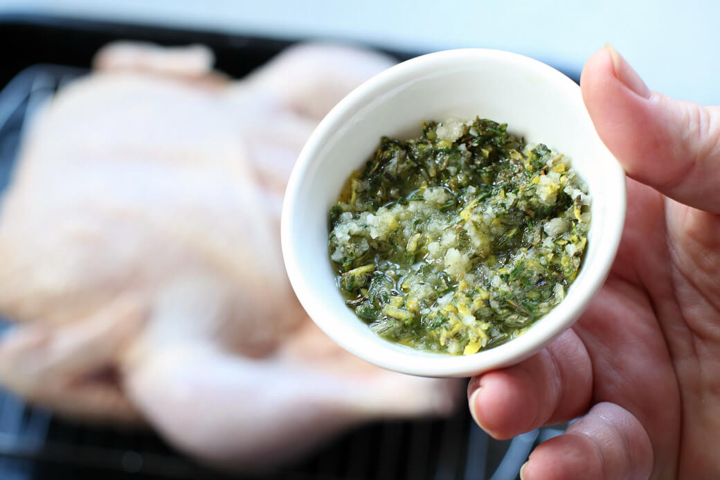 Dry Brine Chicken - A hand holds a small white bowl containing lemon herb dry rub with a raw chicken sitting in the background.