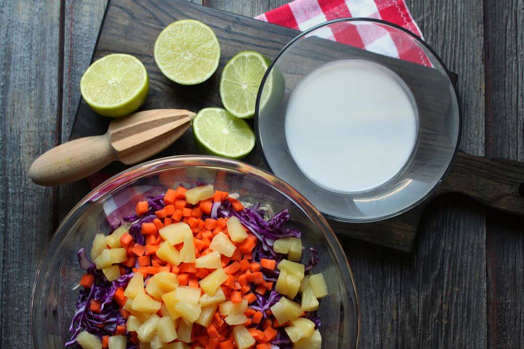 Ingredients for a multicoloured coleslaw with pineapple, carrots, coconut cream, and lime.