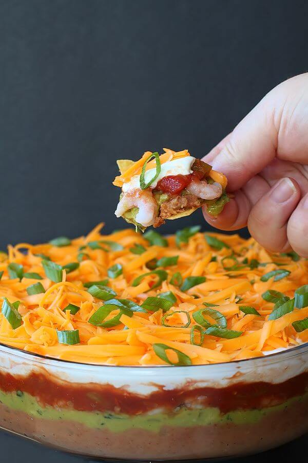 7 Layer Dip - A hand holding a nacho chip topped with layered dip above a glass dish of layered dip.