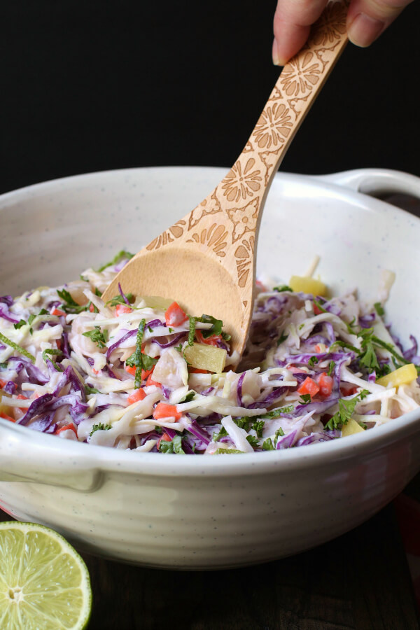 A multicoloured coleslaw with pineapple and lime in a white bowl with a wooden spoon.