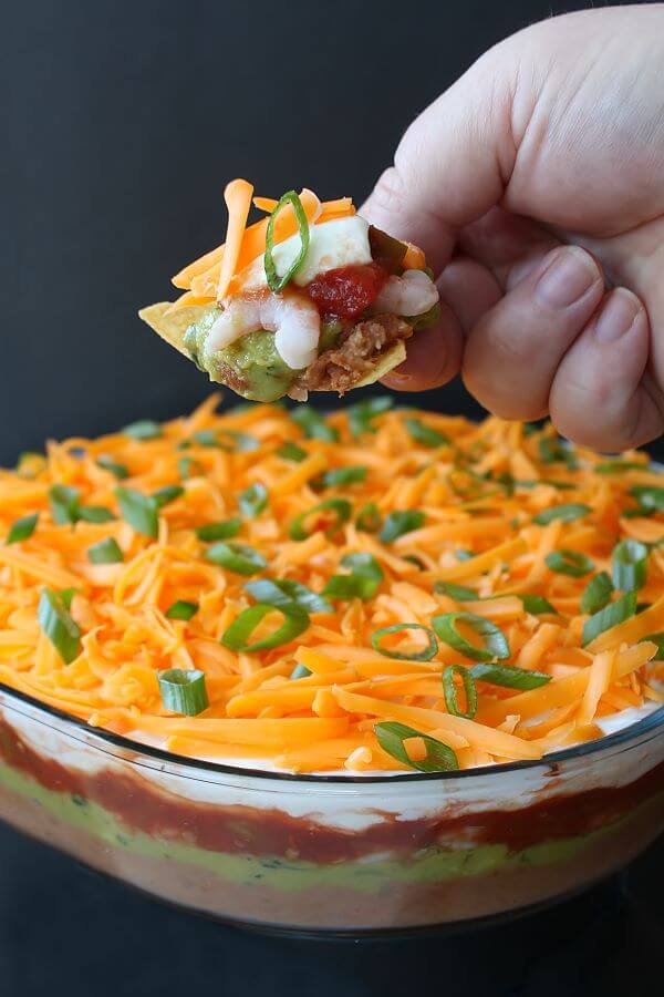 7 Layer Dip - A hand holding a nacho chip topped with layered dip above a glass dish of layered dip.