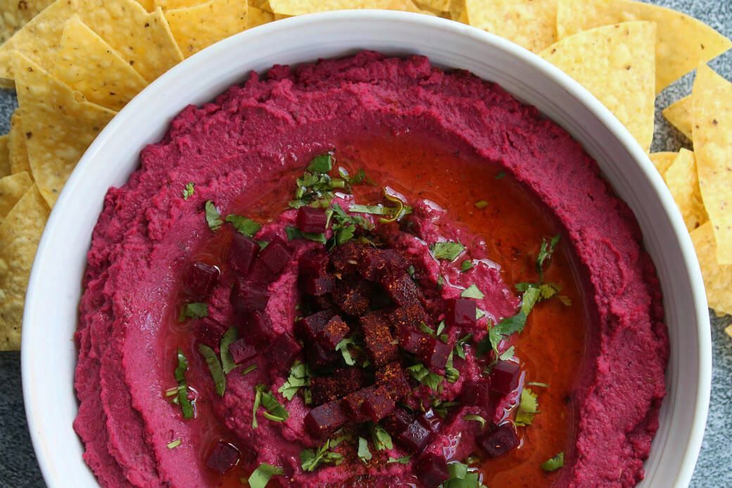 Baharat Roasted Beet Hummus - A bowl of brightly coloured pink hummus garnished with olive oil, beet cubes, and parsley. with a side of chips.