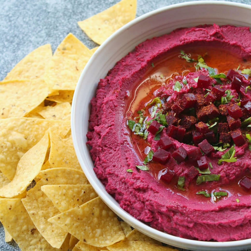 Baharat Roasted Beet Hummus - A bowl of brightly coloured pink hummus garnished with olive oil, beet cubes, and parsley. with a side of corn chips.
