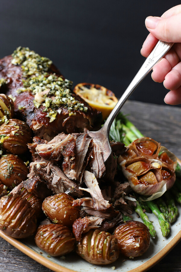 A fork holding pulled lamb meat above a platter filled with a boneless roasted leg of lamb, potatoes, asparagus, roaster garlic, and charred lemon.