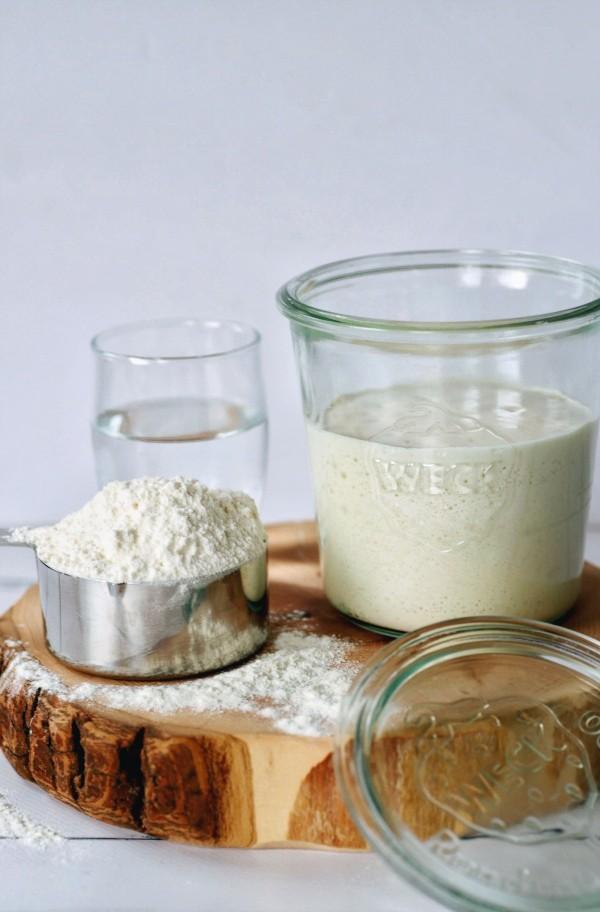 How to Make Sourdough Starter - sourdough starter in a glass jar surrounded by measuring cup of flour and jar of water. 