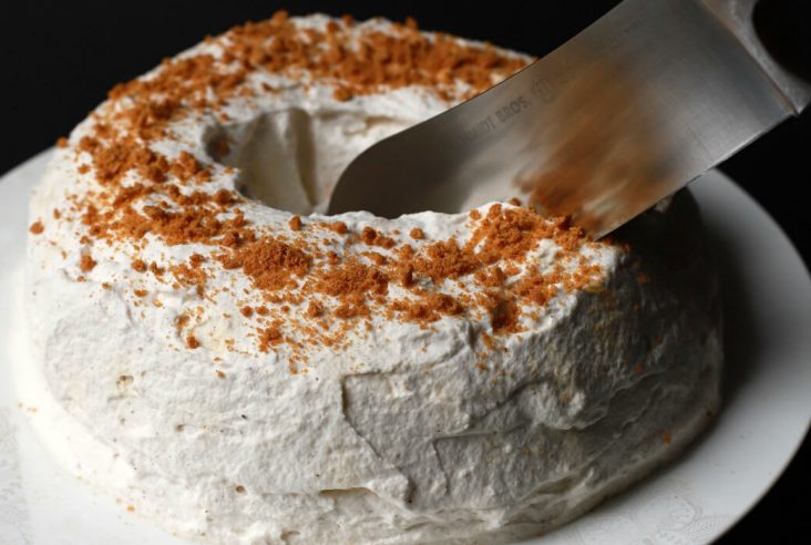 A knife cuts into a white circular cake topped with ginger crumbs.
