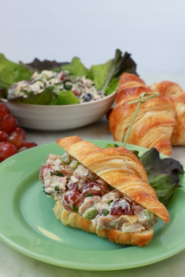 Three croissants, a bunch of red grapes, and a lettuce lined bowl filled with chicken salad. A made up chicken salad croissant sits on a green plate.