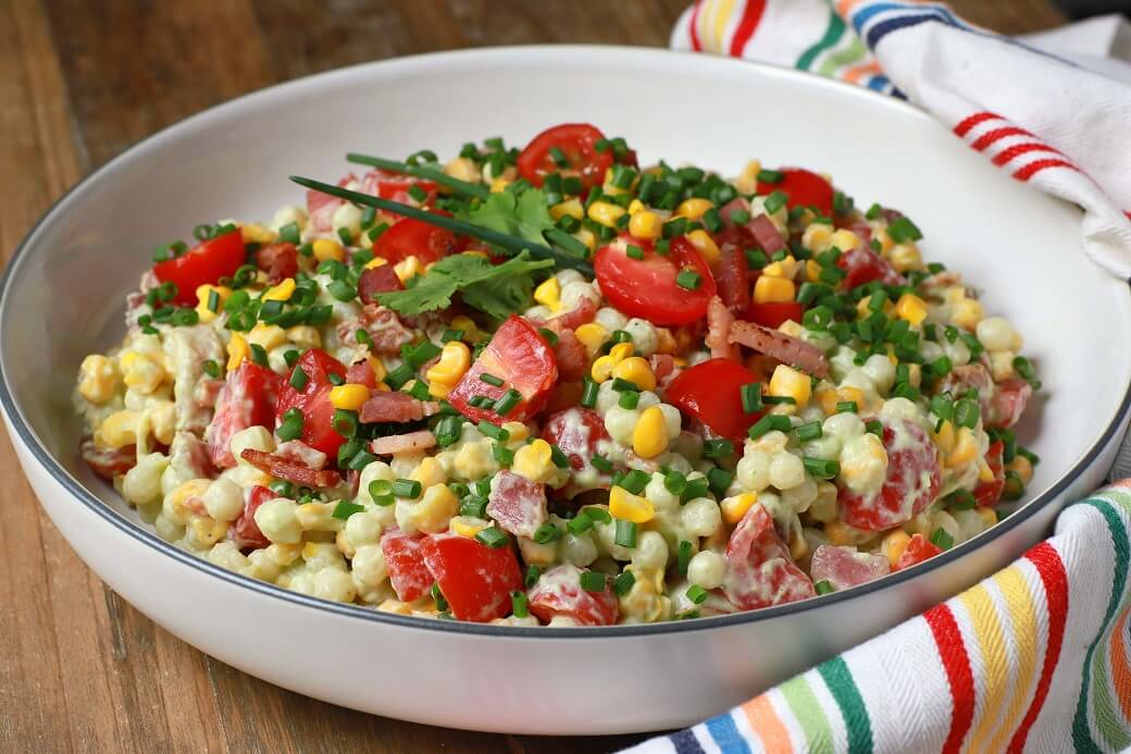 A bright and cheerful salad containing red grape tomatoes, corn, bacon, cilantro, and pearl couscous in a white bowl.