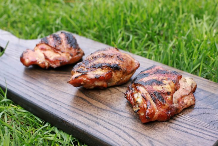 Three perfectly smoked and grilled chicken thighs on dark wooden board in the green grass.