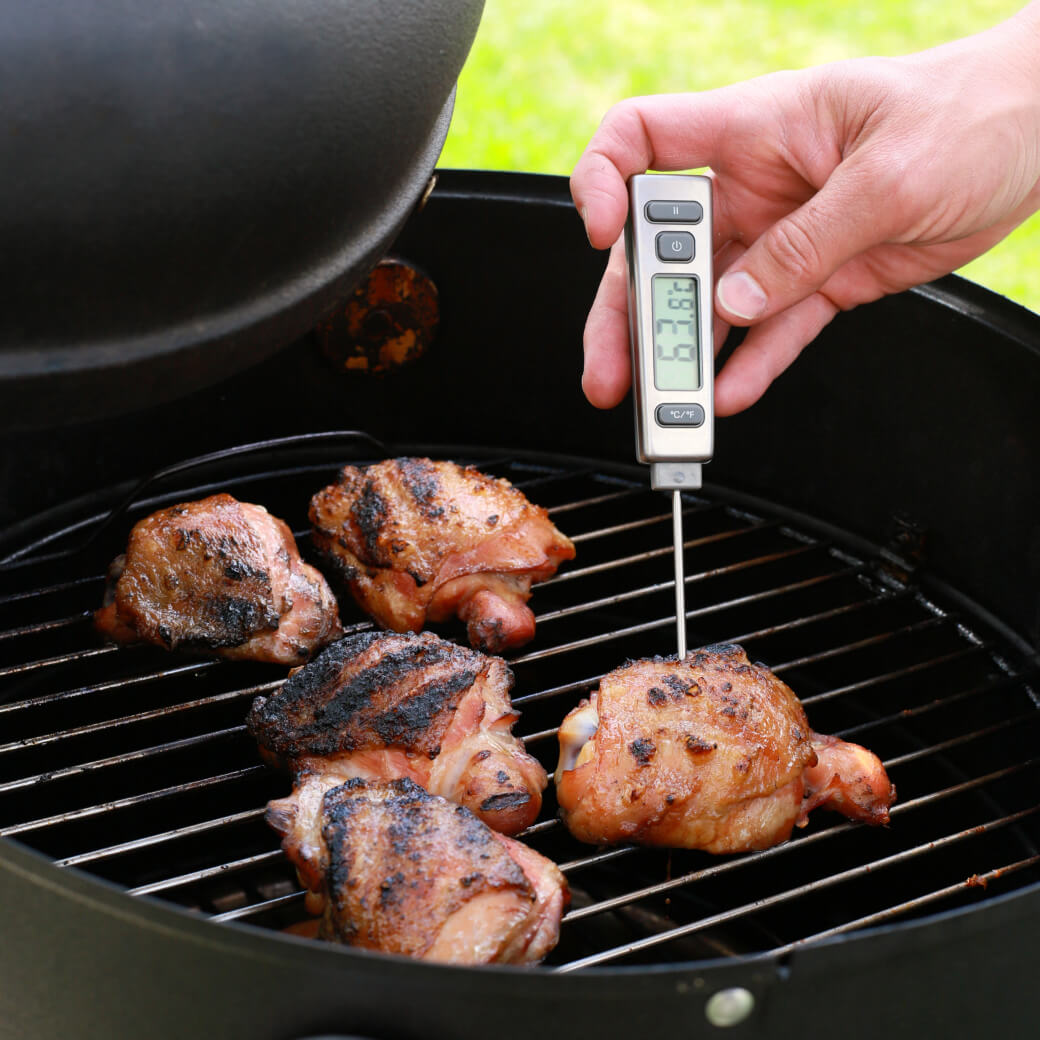 A chef takes the temperature of a chicken thigh on a hot grill using a thermometer.