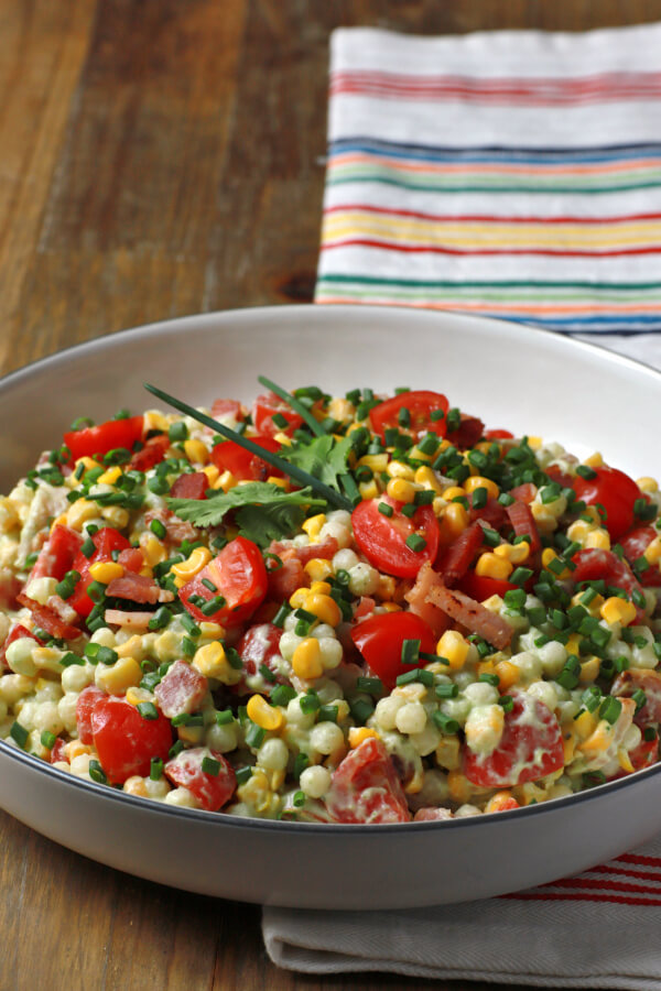 A bright and cheerful salad containing red grape tomatoes, corn, bacon, cilantro, and pearl couscous in a white bowl.