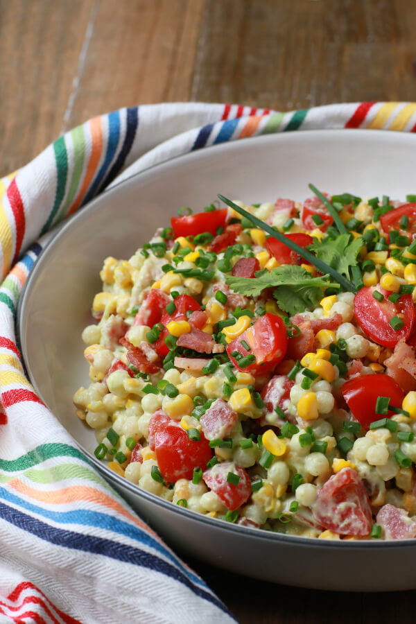 Smoked Corn Bacon Avocado Pasta Salad is a bright and cheerful salad containing red grape tomatoes, corn, bacon, cilantro, and pearl couscous in a white bowl.