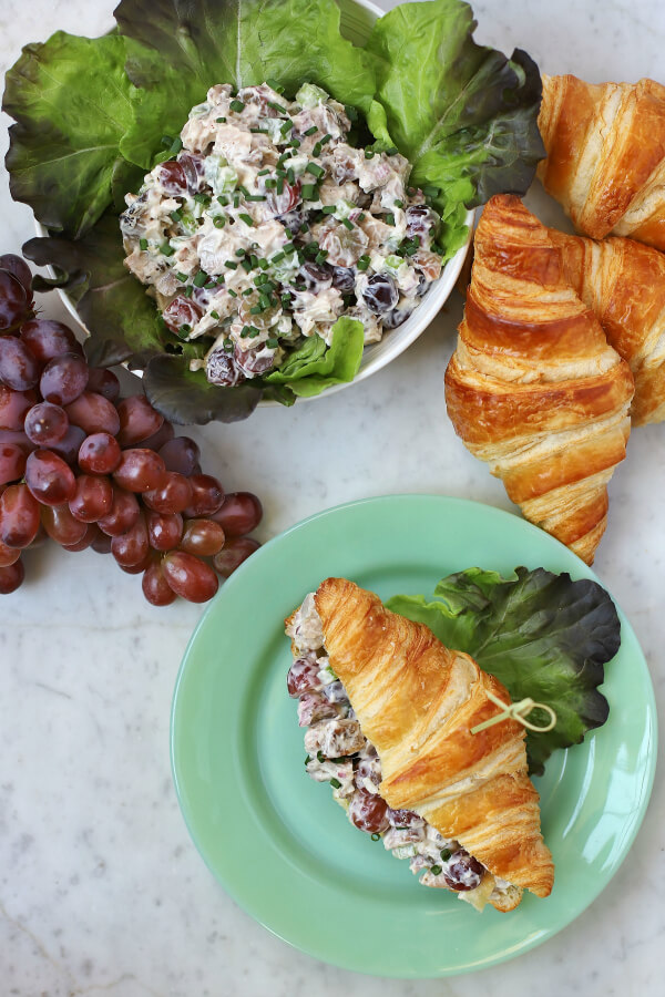 Three croissants, a bunch of red grapes, and a lettuce lined bowl filled with chicken salad. A made up chicken salad croissant sits on a green plate.