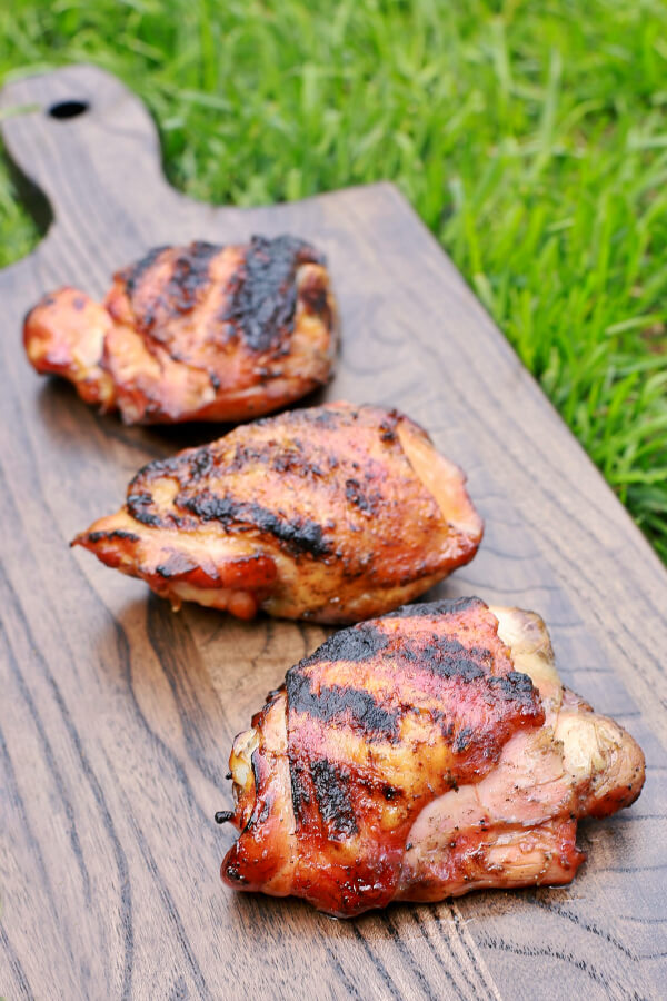 Three perfectly smoked and grilled chicken thighs on dark wooden board in the green grass.