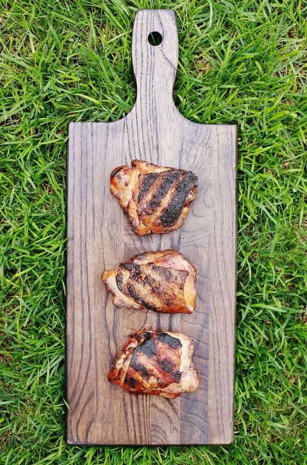 Three perfectly smoked and grilled chicken thighs on dark wooden board in the green grass.