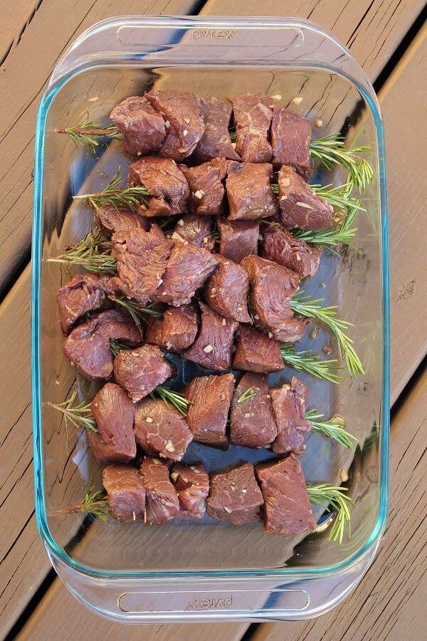 Pieces of raw beef marinating on rosemary skewers in a glass dish.