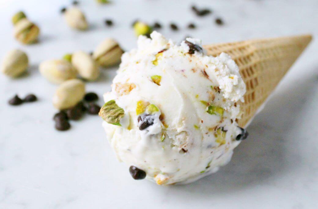 A scoop of pistachio and chocolate chip studded ice cream in a cone lying on white marble.