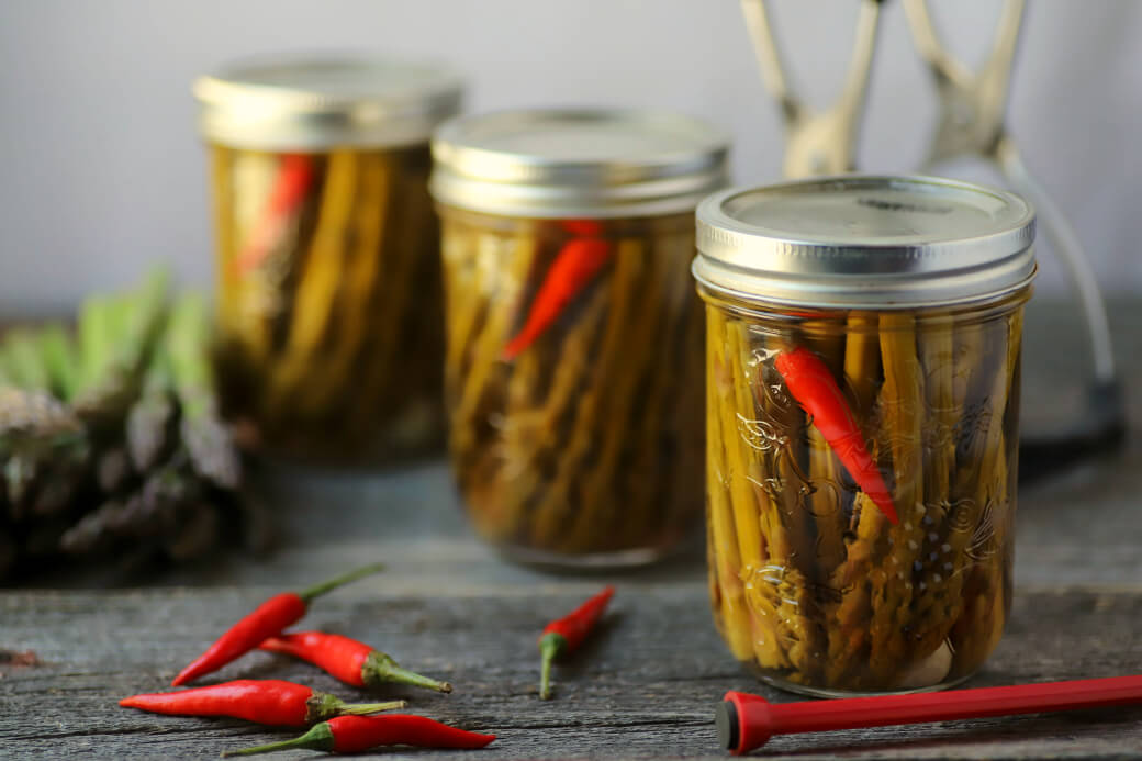 Two clear glass jars filled with green asparagus, a garlic clove, and a red hot pepper after processing in a hot water bath.