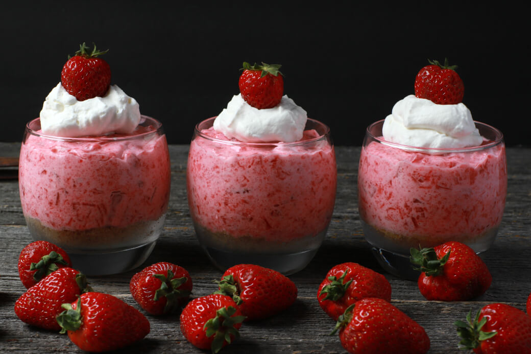 Three pink rhubarb strawberry jello desserts in cups with strawberries and cream.