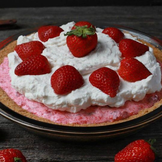 A pink rhubarb strawberry pie in a graham crust topped with strawberries and whipped cream.