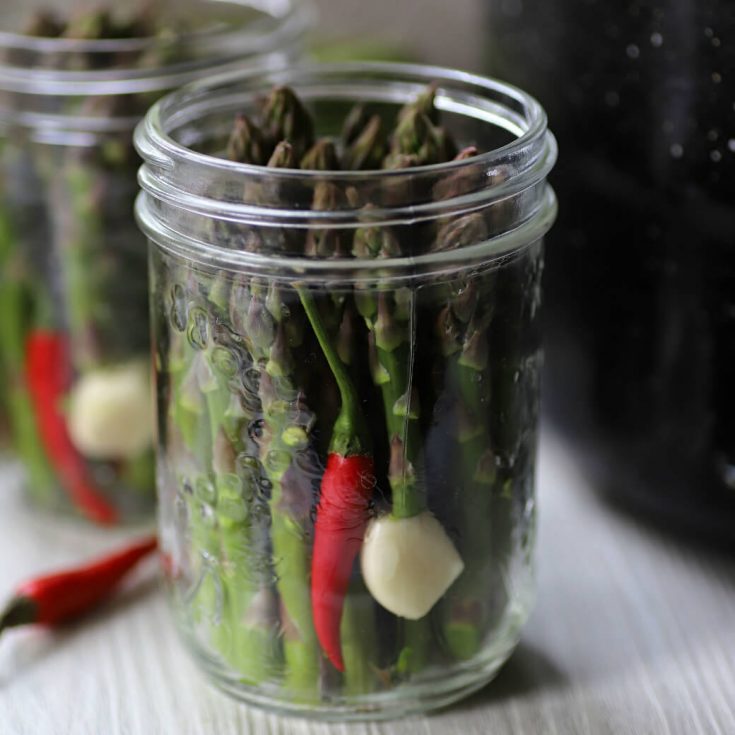 A clear glass jar filled with green asparagus, a garlic clove, and a red hot pepper.