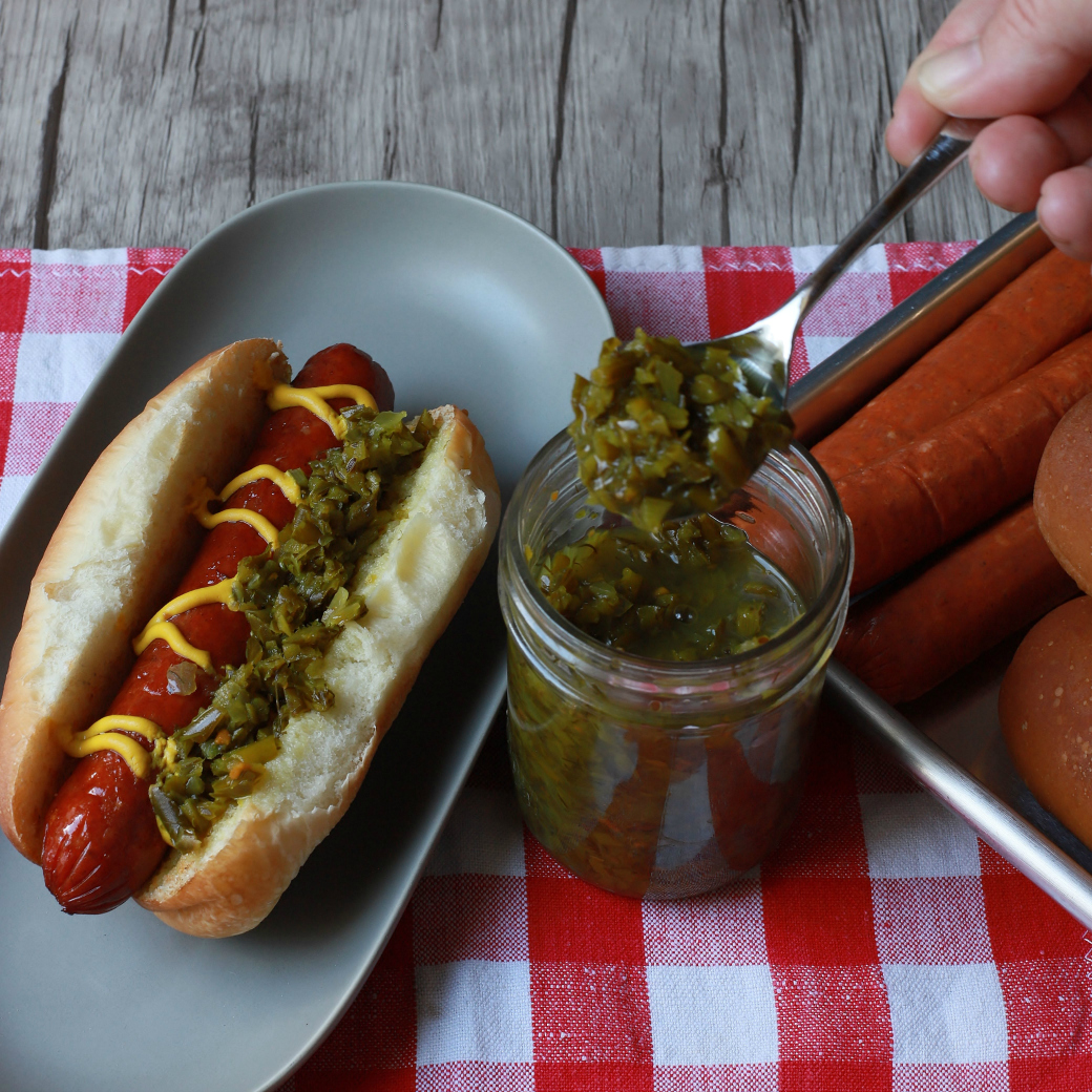 A jar of Garlic Scape Relish next to a loaded hot dog.