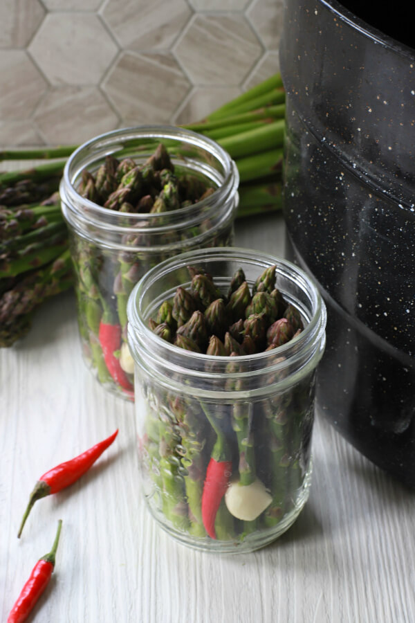 Two clear glass jars filled with green asparagus, a garlic clove, and a red hot pepper.
