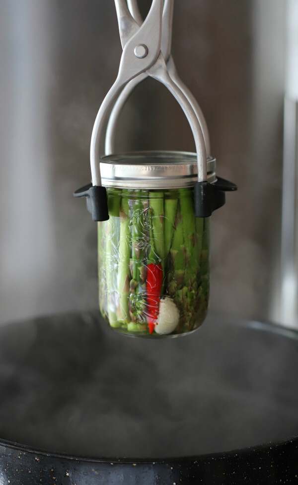 A clear glass jar filled with green asparagus, a garlic clove, and a red hot pepper being lowered into a hot water canner.