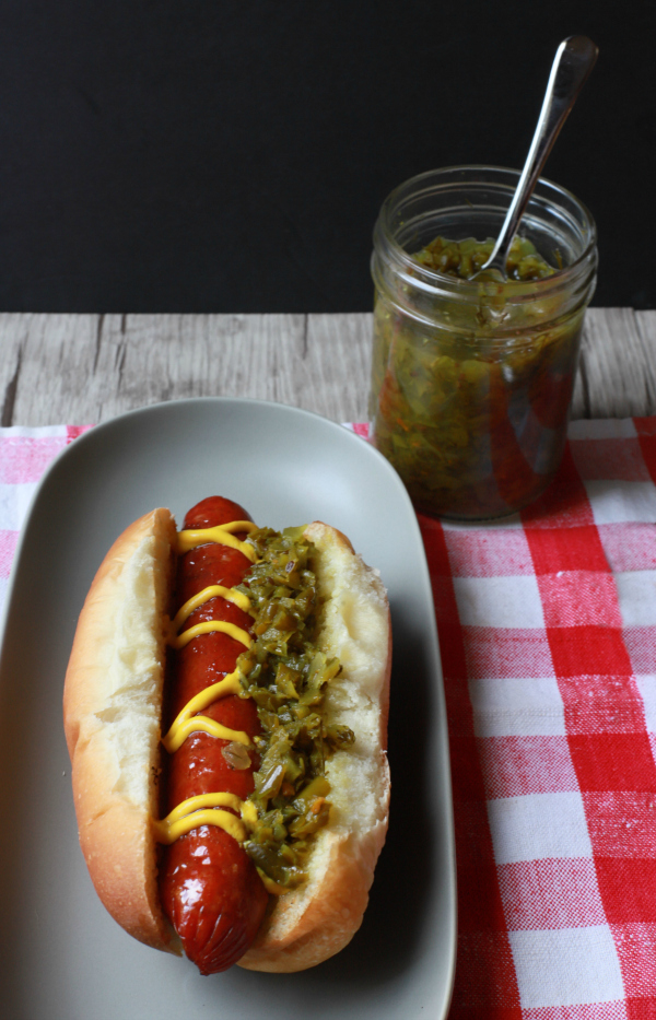 A jar of Garlic Scape Relish next to a loaded hot dog.