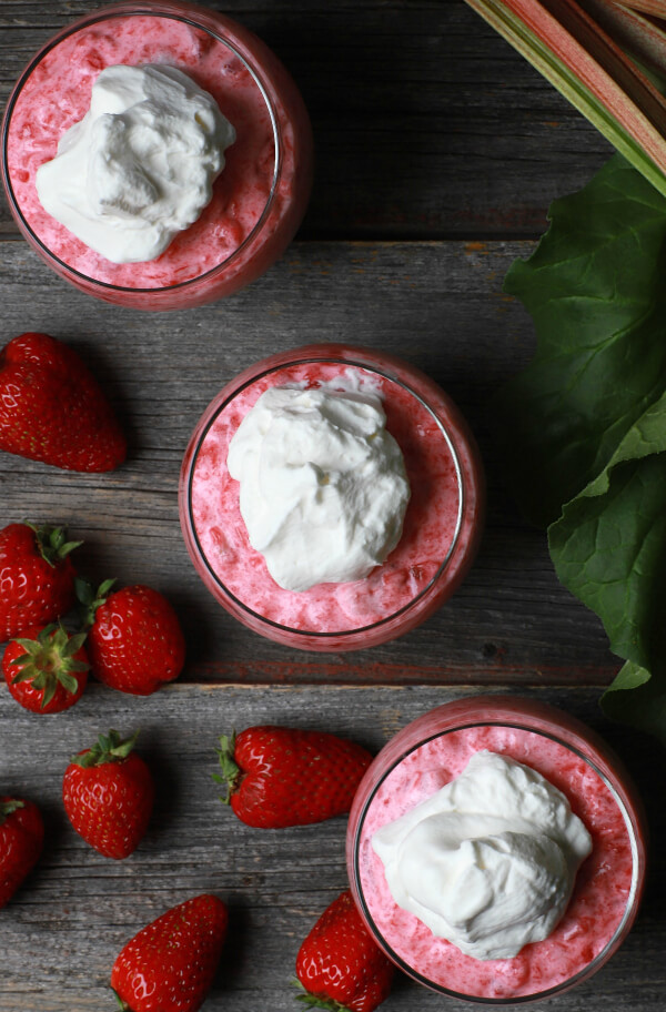 Three pink rhubarb strawberry jello desserts in cups with strawberries and cream.