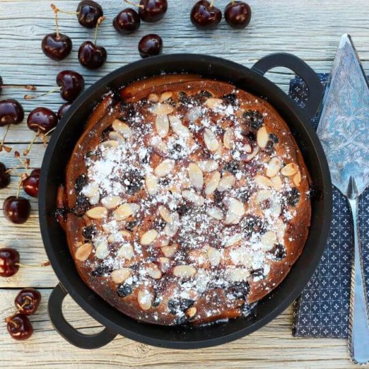 A cast iron pan containing baked cherry clafoutis topped with almonds and powdered sugar surrounded by fresh cherries.
