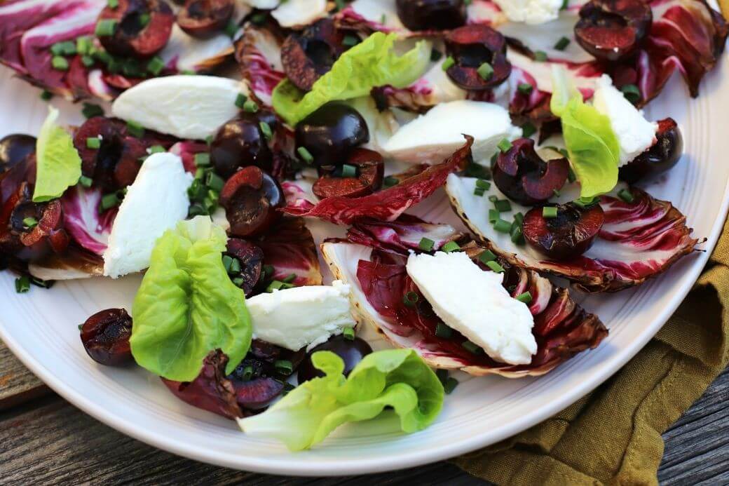 A beautiful salad of grilled radicchio, bright red cherries, green lettuce, and white goat cheese quenelles on a white plate.