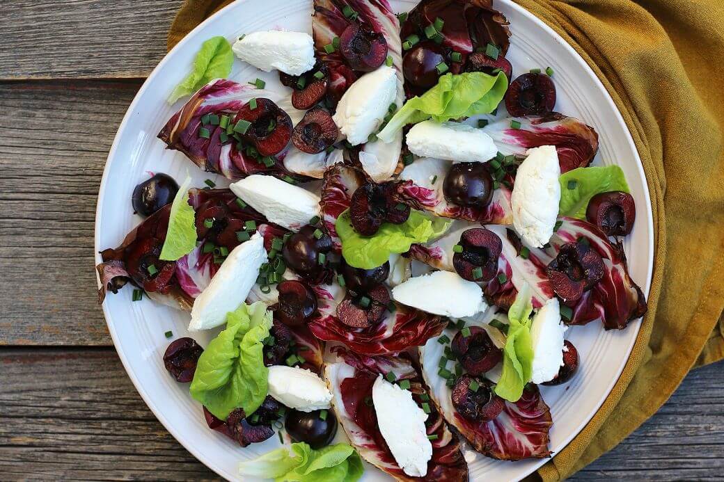 A beautiful salad of grilled radicchio, bright red cherries, green lettuce, and white goat cheese quenelles on a white plate. 