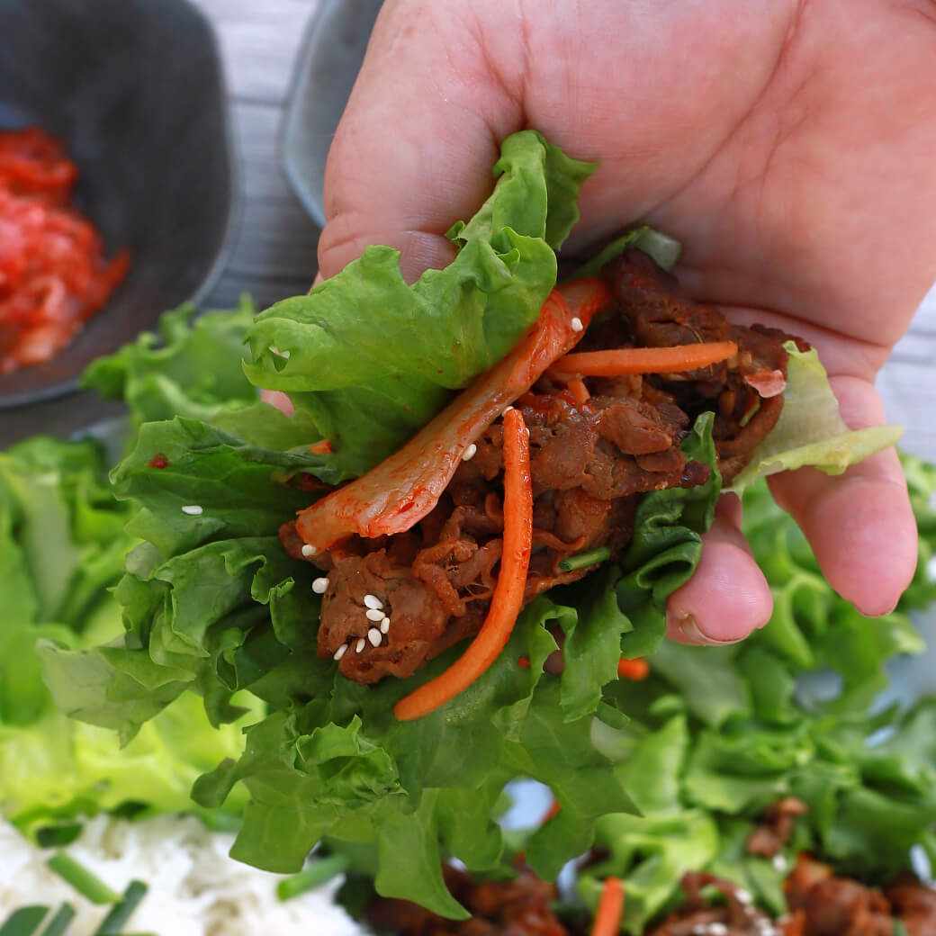 A hand holds a lettuce wrap filled with caramelized beef, onions, carrots, and sesame seeds.